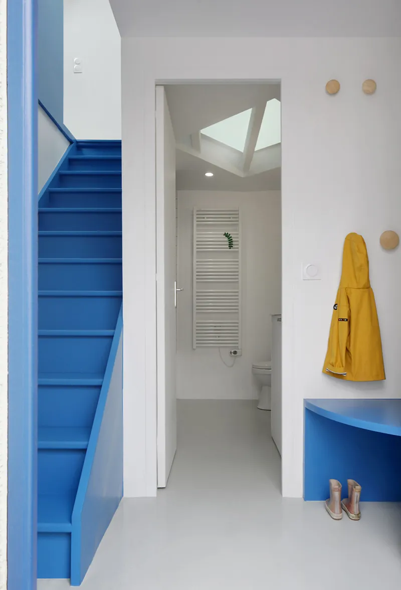 White corridor connecting blue stair and bathroom with skylight beyond yellow raincoat and wall hooks