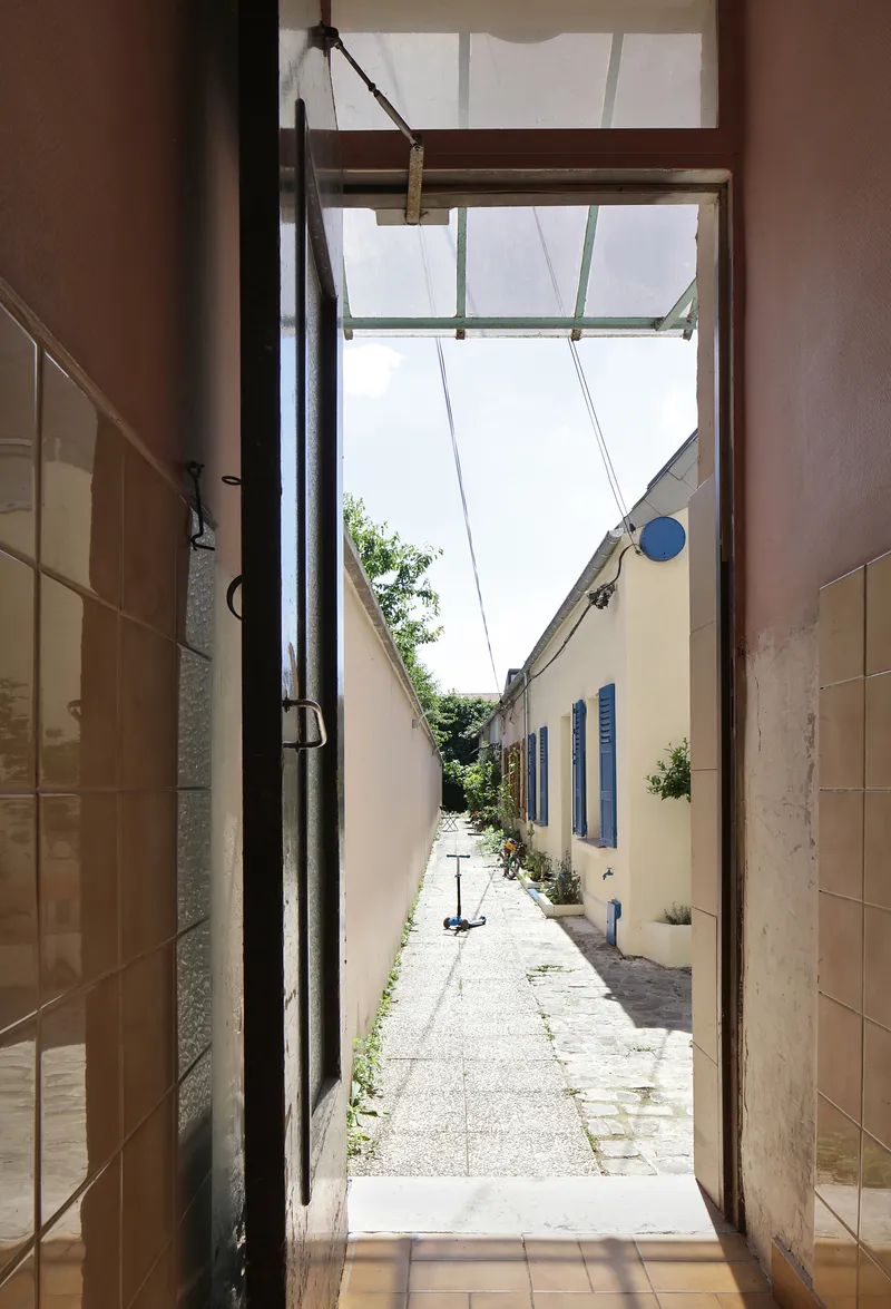 View through a brown-tiled threshold toward a narrow paved lane with blue shutters and greenery