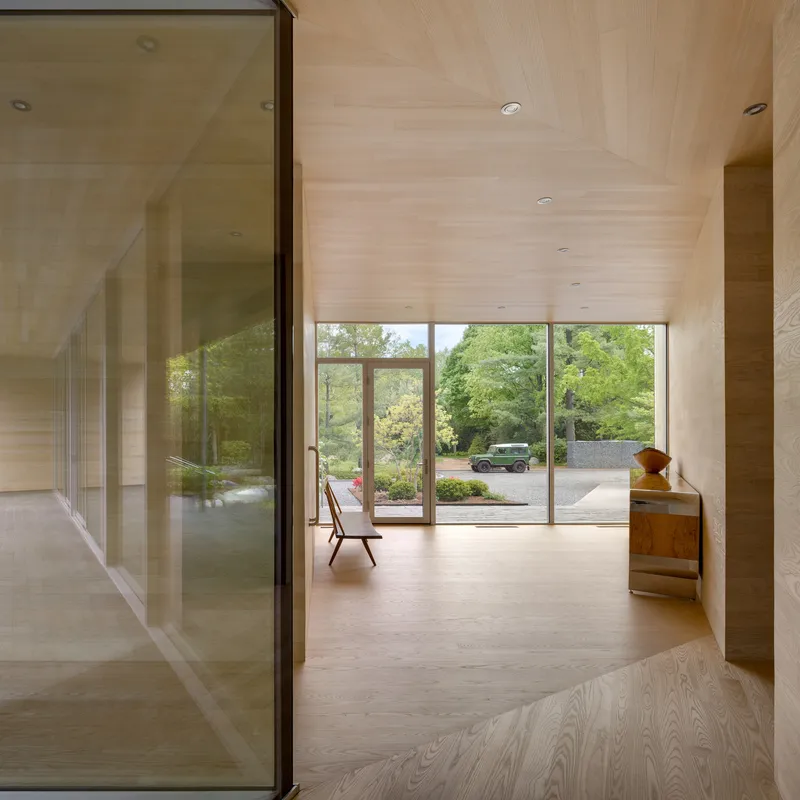 Wood-lined corridor with glass partition reflecting view through glazed doors to landscaped driveway