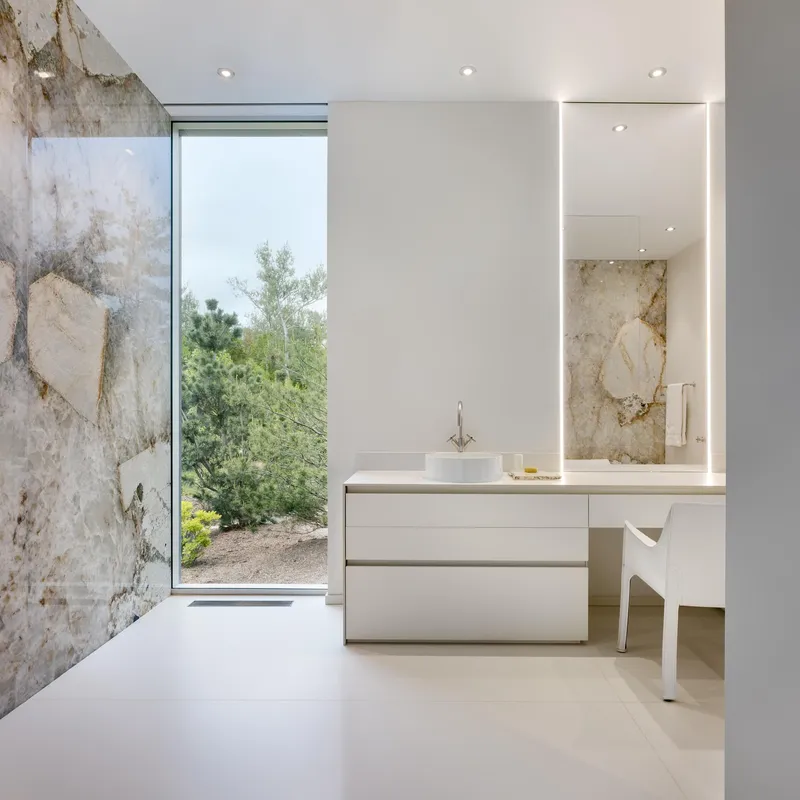 Bathroom with weathered stone wall, floor-to-ceiling glazing overlooking green landscape and white vanity with backlit mirror