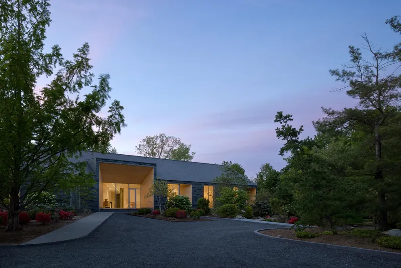 Single-storey residence with slate-clad facade and cantilevered entry canopy at dusk, surrounded by planted beds and trees