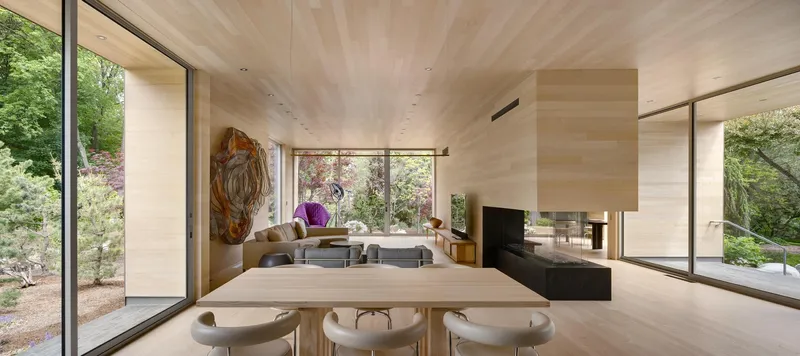 Open living and dining space with plywood ceiling and floor-to-ceiling glazing overlooking the garden