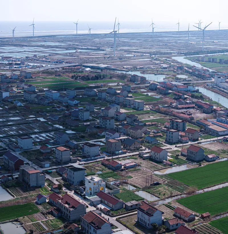 Aerial view of a low-rise residential settlement with agricultural plots and wind turbines on the horizon