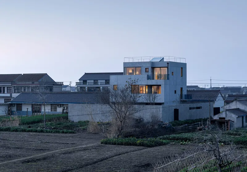 White concrete house with rooftop terrace rising above neighboring buildings and cultivated fields at dusk
