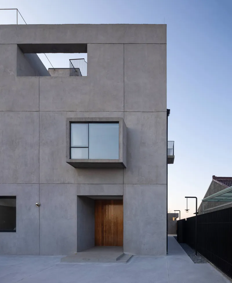 Street-facing concrete facade with timber entry door and projecting box window under a clear sky