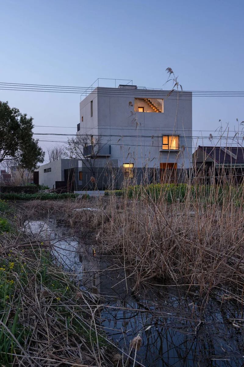 Concrete facade with lit windows at dusk across a narrow irrigation channel with dry winter grasses