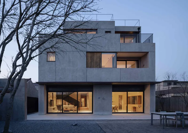 Rear elevation with large glazed openings and cantilevered terraces against a gravel courtyard at twilight