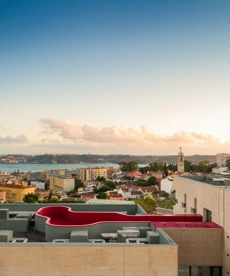 Rooftop view of a red curved volume atop a buff brick facade overlooking the harbor at sunset