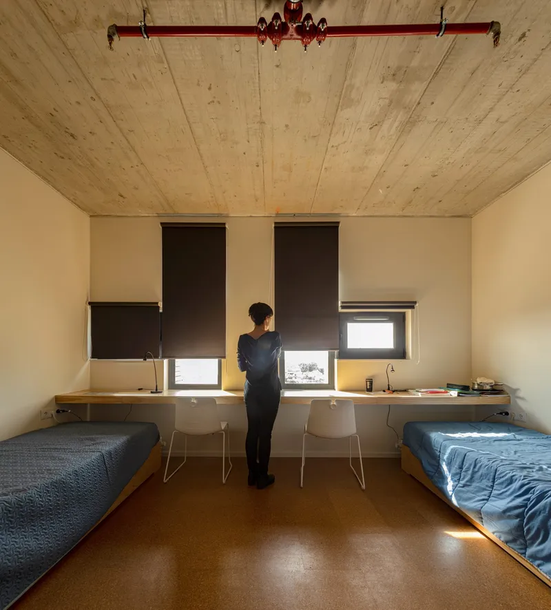 Shared bedroom with two beds and floating desk beneath partially drawn roller shades with a person standing