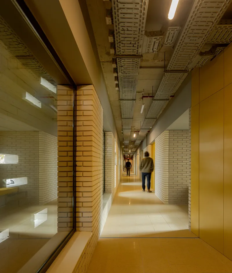 Long corridor with cream brick walls and exposed ceiling as two people walk beneath overhead lighting