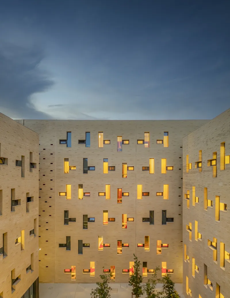 Interior courtyard surrounded by brick walls with illuminated staggered windows and small trees at twilight