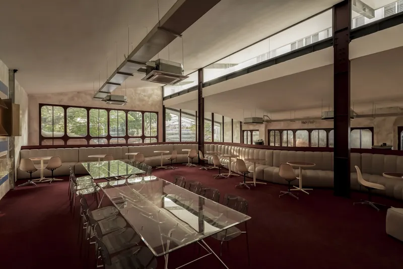Long transparent communal table with acrylic chairs on burgundy carpet flanked by curved banquettes and structural columns