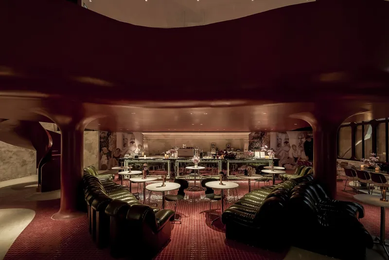 Interior lounge with curved burgundy ceiling, tufted seating and round tables illuminated by warm pendant lights