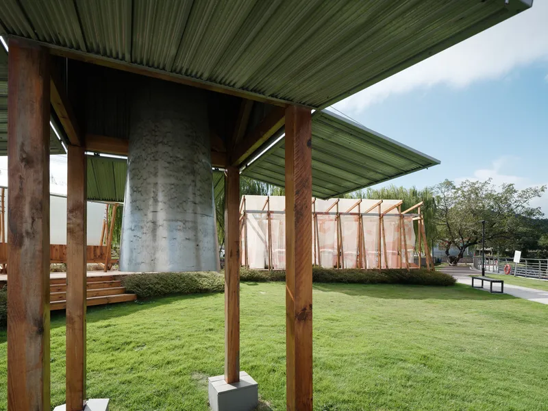 Sheltered pavilion with corrugated metal roof, timber posts, and concrete bell framed against the lawn and surrounding structures