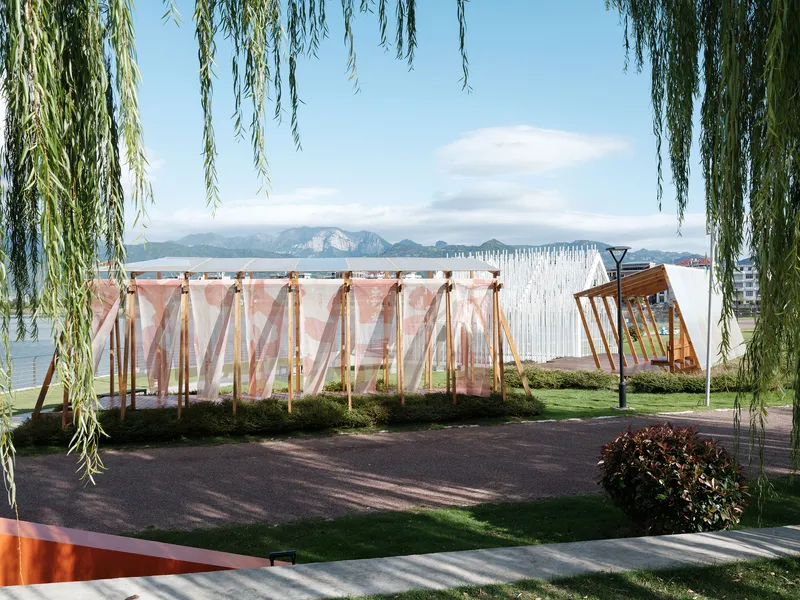 Timber frame pavilion with pink translucent fabric panels framed by willow branches along a lakeside path