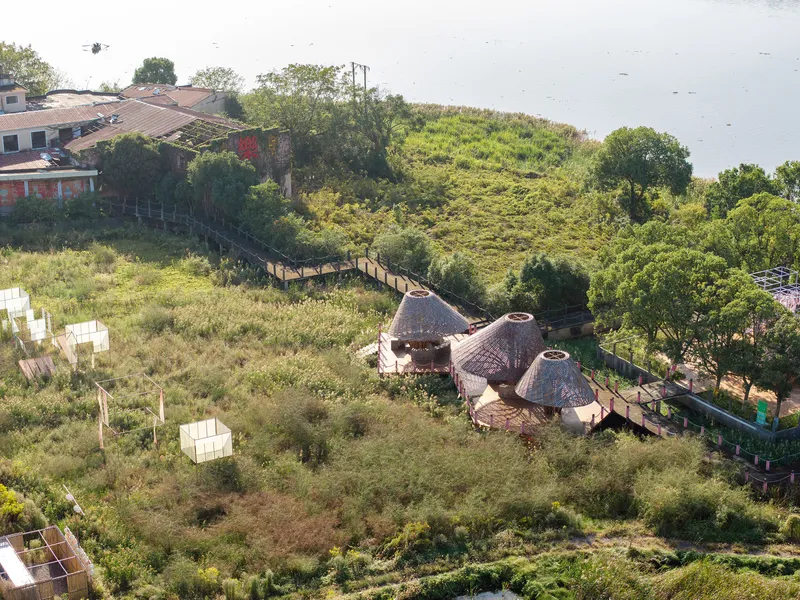 Aerial view of three conical bamboo roofs linked by timber walkways through a wetland landscape