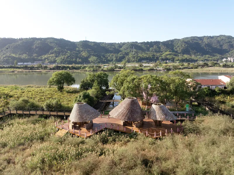 Three thatched roof structures connected by timber decking platforms overlooking a river and distant mountains