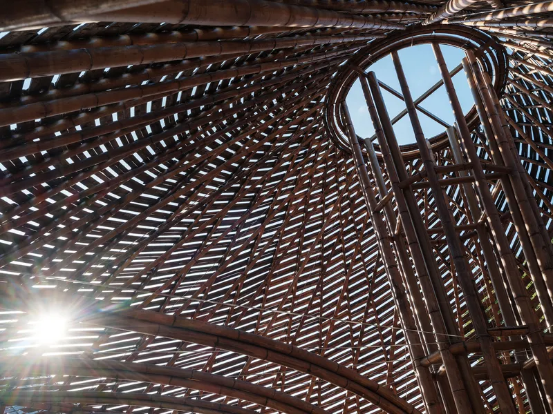 Interior view looking up through a woven bamboo roof structure with sunlight filtering through gaps