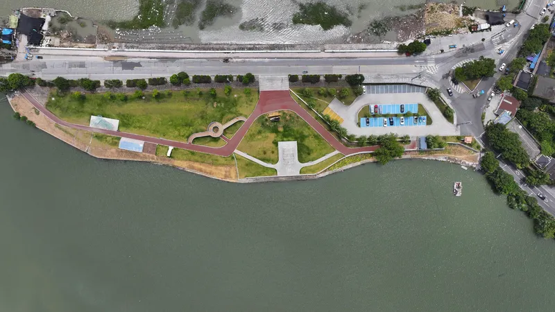 Aerial view of the waterfront park with curving red pathways, lawn areas, and white vertical-slatted pavilions along the shore