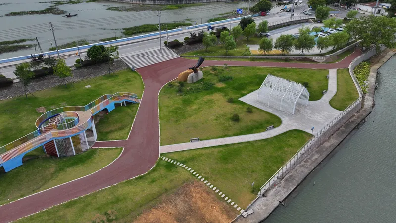 Drone view showing circular observation platform, timber pavilions, and red walking paths threading through the green riverside park