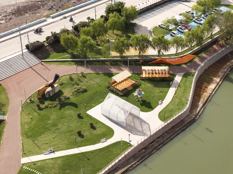 Aerial perspective of waterside installations including timber-clad structures and white vertical-screen pavilion beside the paved path
