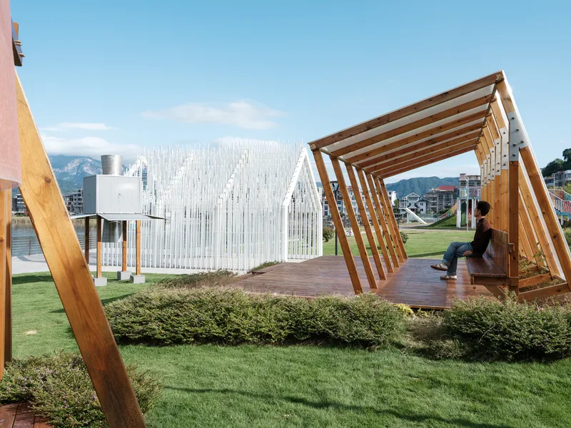 Timber-framed triangular seating shelter with wooden deck, planted bed, and white slatted pavilion in the background