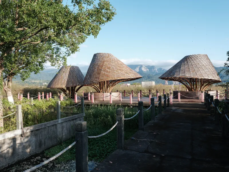 Three thatched bamboo canopies with branching timber columns along a boardwalk with distant mountains visible