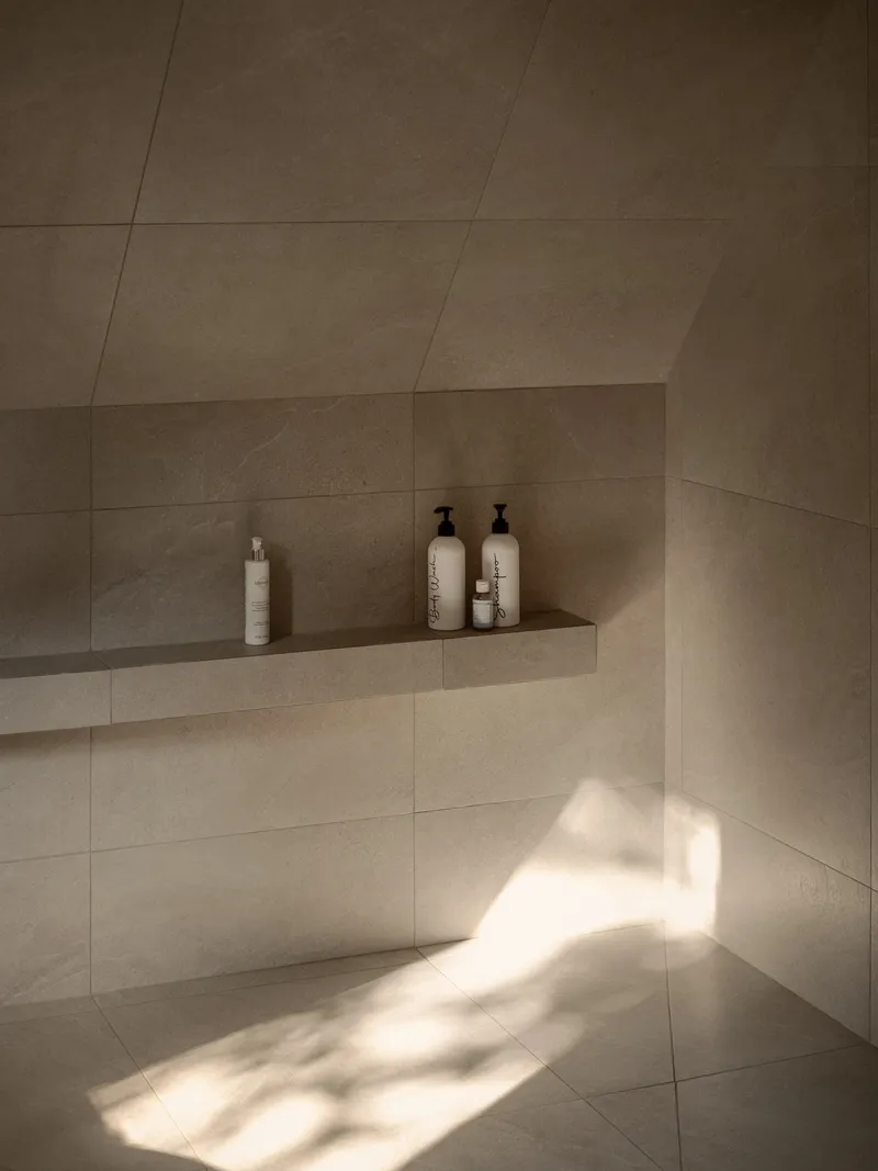 Recessed stone tile shower niche with white bottles illuminated by sunlight filtering across the floor