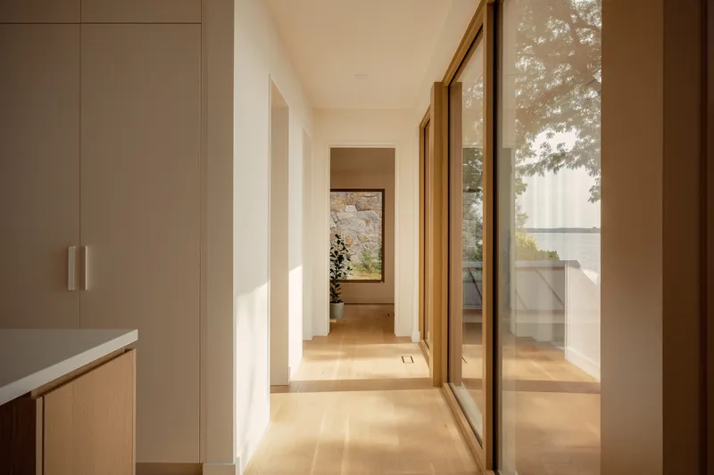 Hallway with full-height glazing and timber frames overlooking waterside terrace in afternoon light