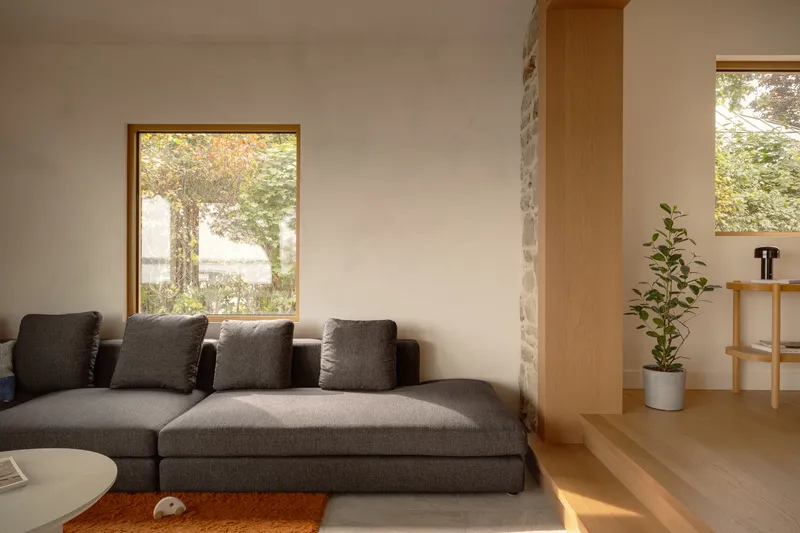 Living room with grey sectional sofa and timber-framed window overlooking trees in autumn light