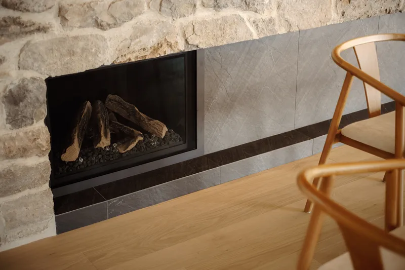 Gas fireplace insert framed by grey stone tiles set into a sandstone wall beside a timber chair