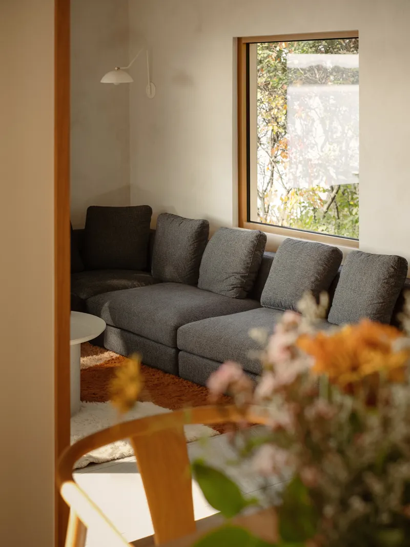 Timber framed window with drawn shade overlooking a sofa in soft daylight with flowers in foreground
