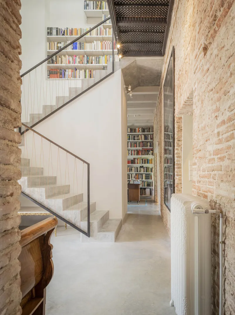 Concrete staircase with steel railings ascending between exposed brick walls and floor-to-ceiling bookshelves
