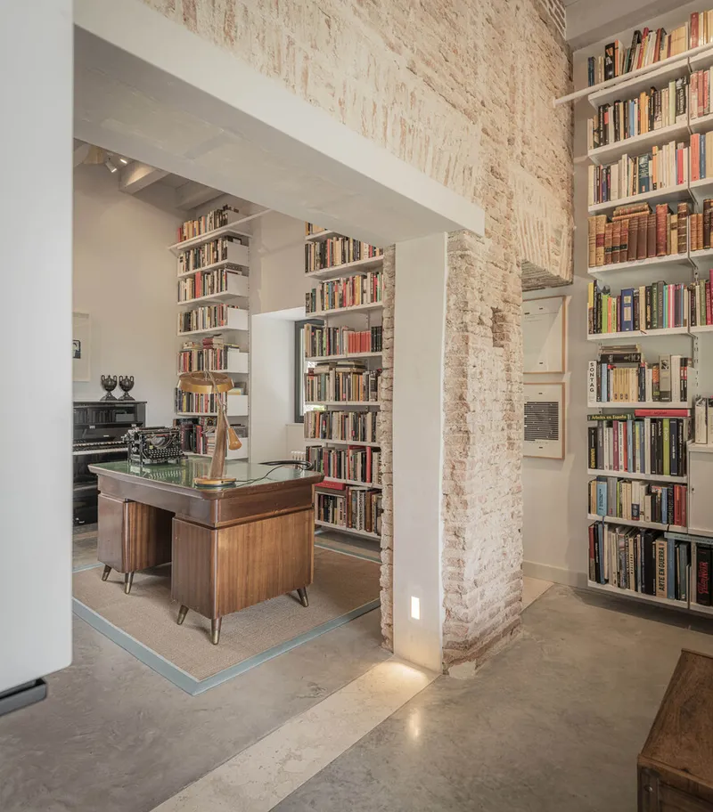 Interior view through exposed brick columns framing a vintage desk and floor-to-ceiling bookshelves on concrete floors