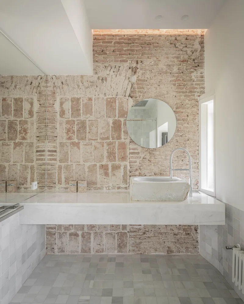 Bathroom with floating marble vanity and vessel sink against patchwork brick and plaster wall under skylight