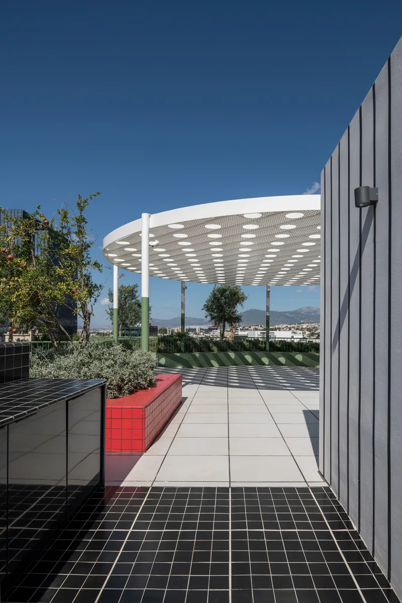 Rooftop terrace with circular perforated canopy, black tile paving, and red bench under clear blue sky
