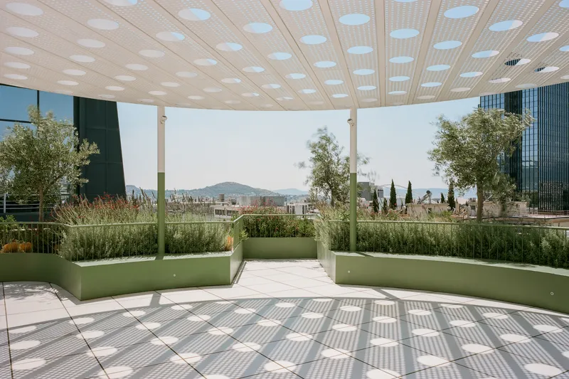 Circular canopy with perforated ceiling above green planted beds and tiled terrace overlooking distant hills