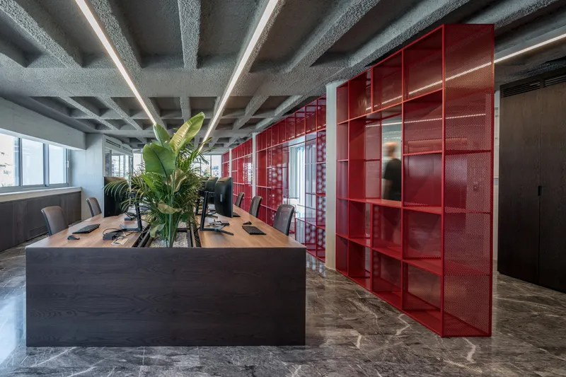 Office interior with exposed concrete ceiling, red perforated steel partitions, and a figure passing between workspaces