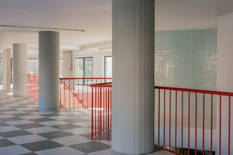 Upper floor circulation space with red tubular steel railings, round concrete columns, and checkered tile flooring