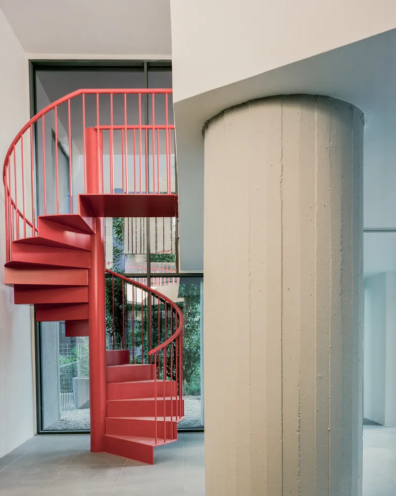 Red spiral staircase beside a cylindrical concrete column with garden views through glazed opening