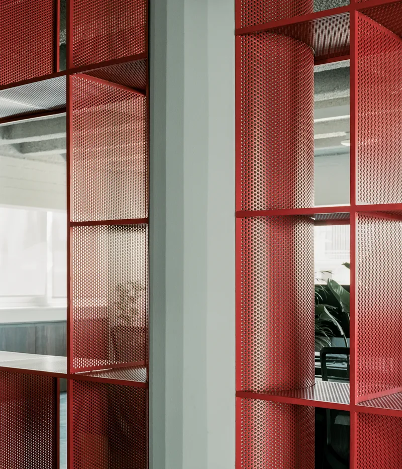 Close-up of red perforated metal shelving units with curved segments reflecting light from workspace beyond