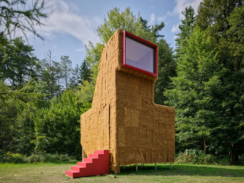 Textured straw bale facade with red staircase and upper window framed against deciduous trees