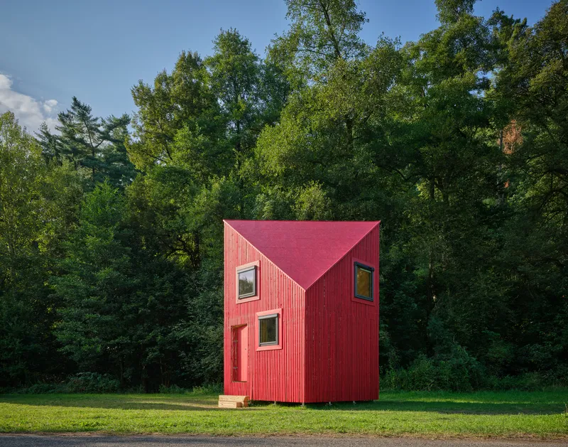 Red corrugated metal cabin with gable roof and asymmetrical windows in a forest clearing