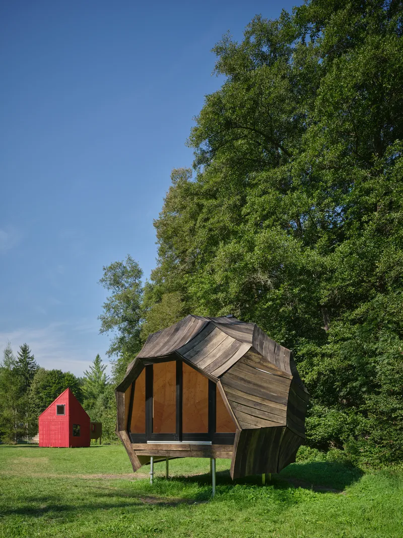 Faceted timber pavilion on stilts with angular roof planes beneath tall deciduous trees