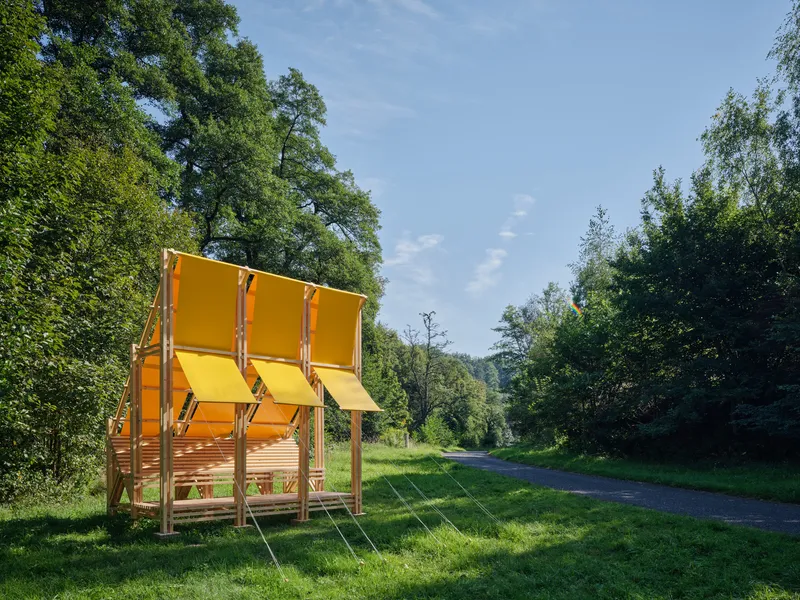 Yellow canopy structure with rope suspension and timber slat base along a rural road
