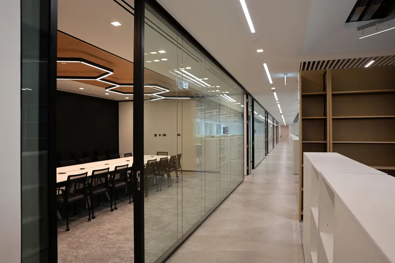 Glass-walled corridor with conference rooms on one side and timber shelving opposite under linear ceiling lights