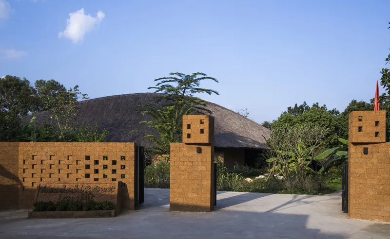 Entry gate with perforated rammed earth walls and timber towers flanking a concrete path under a blue sky