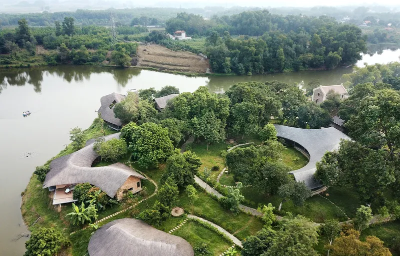 Drone view of the compound showing thatched pavilions and pathways organized around mature trees by the river