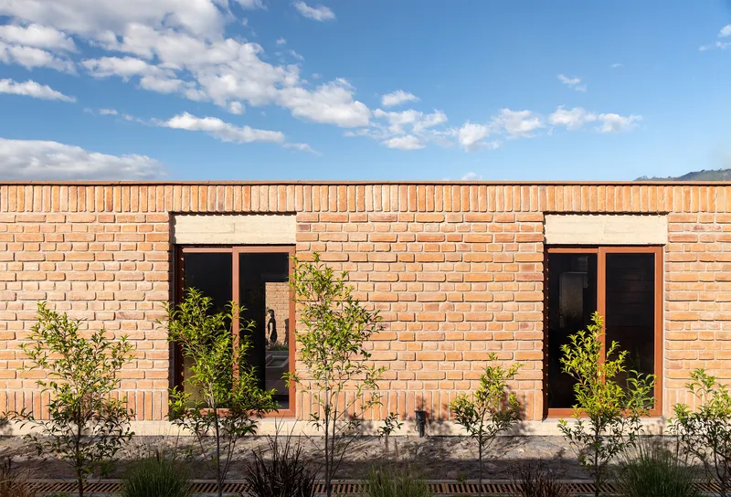 Brick facade with two symmetrical window openings and young trees planted along a gravel base