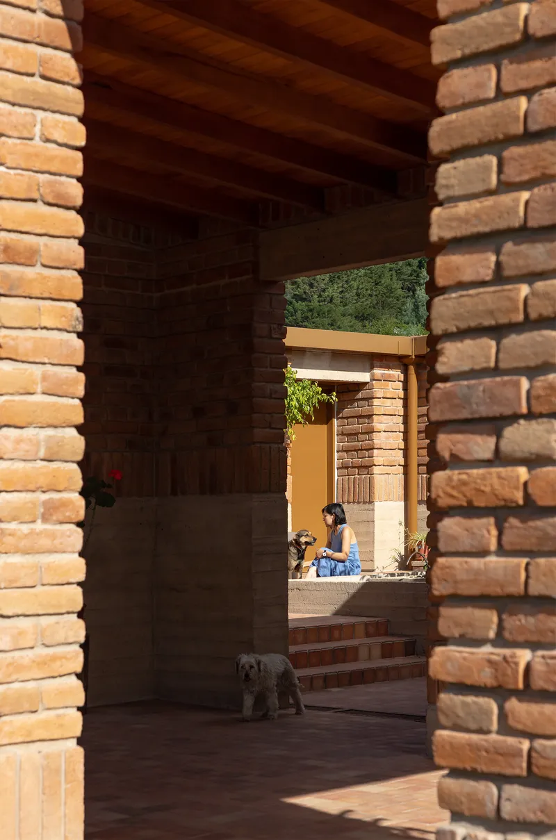 Shaded brick corridor with timber ceiling beams framing a view of a seated figure and dog